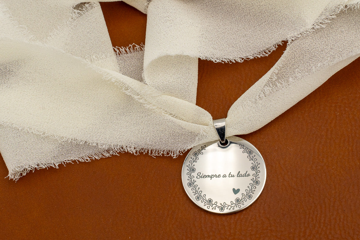 Anhänger aus spiegelpoliertem Edelstahl mit dem Spruch „Immer an deiner Seite“ und Blumenrand, an einem elfenbeinfarbenen Chiffonband