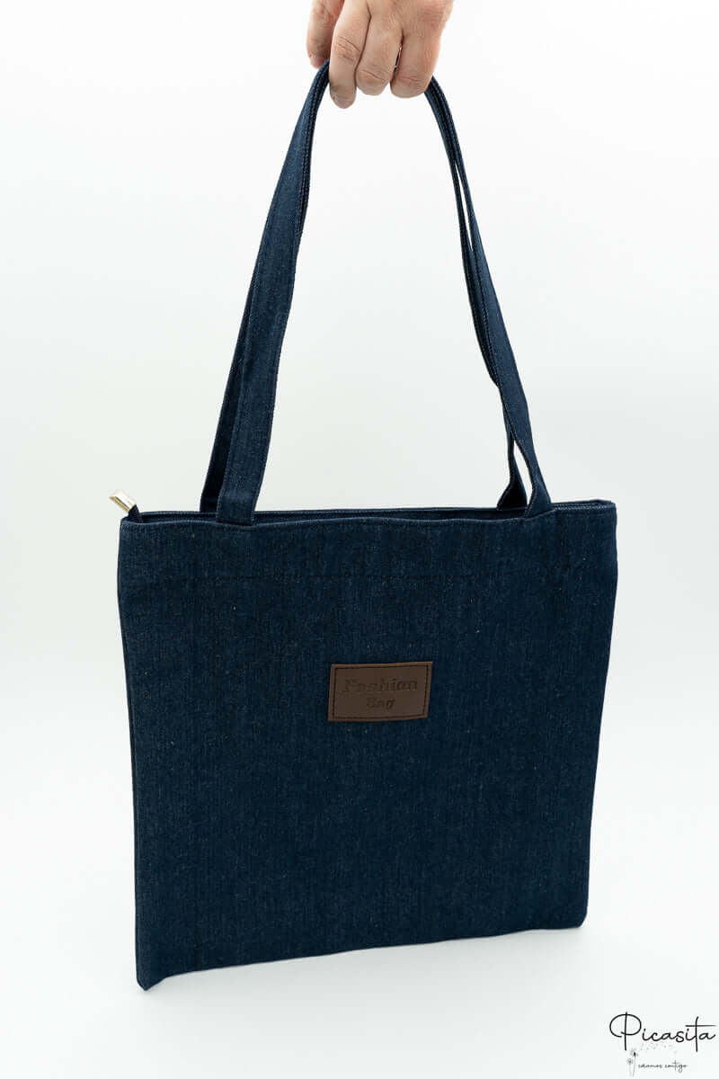 Bolso vaquero tipo tote sostenido por la mano con parche central que indica Fashion Bag.